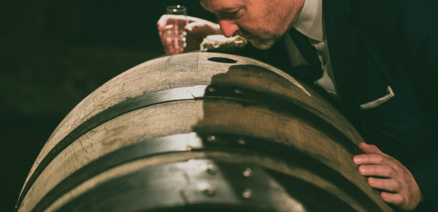 Man holding a glass of whisky looking into a barrel with his hand resting on it
